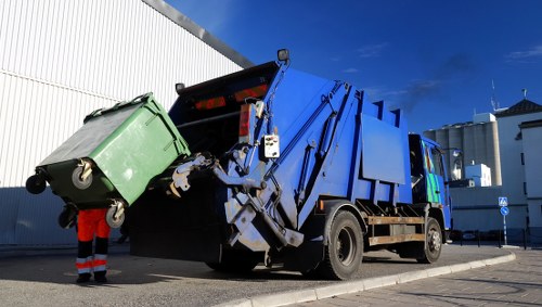 A large blue waste collection truck with a mechanical compactor at the rear is in the process of collecting rubbish from a green wheeled bin, which is tilted and partially lifted into the truck's hopper. The bin is made of plastic with a textured surface and is positioned on a paved area adjacent to a white industrial building or warehouse. A worker wearing an orange high-visibility jacket and red trousers is standing nearby, guiding the bin. The scene is set outdoors under a clear blue sky with a few clouds, and sunlight casts shadows on the ground, indicating daytime. The image depicts a professional waste removal service likely operating within Bayswater, with the focus on the collection of general waste or rubbish as part of commercial waste management, consistent with services offered by Commercial Waste Removal Bayswater.