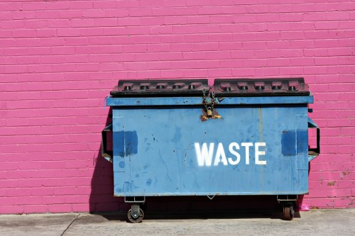 A large, industrial blue waste skip with a hinged lid and a metal latch is positioned on a sidewalk against a pink brick wall. The skip has visible signs of use, including dirt and scratches on its surface, and is mounted on four wheels for mobility. The front of the container prominently displays the word 'WASTE' in white capital letters. The surrounding environment features a concrete pavement, and the shadow cast by the skip indicates daylight. The pink wall in the background provides a contrasting backdrop, suggesting an urban or commercial area, potentially in or near Bayswater. The image reflects a typical scene associated with rubbish collection and waste disposal services offered by companies like Commercial Waste Removal Bayswater, highlighting the practical aspect of waste management in the local area.