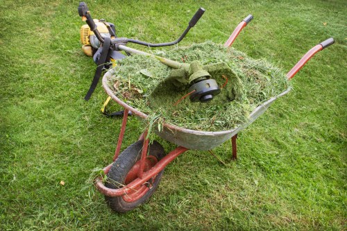 A rusty, red metal wheelbarrow filled with freshly cut green grass and plant debris, positioned on a well-maintained grassy area. The wheelbarrow has a black rubber wheel at the front and two metal handles at the rear, with signs of rust and wear on its surface. Resting atop the grass within the wheelbarrow is a green string trimmer, with a yellow engine casing and a black control handle, partially covered in grass clippings. The background shows more lush green grass, indicating an outdoor garden or lawn setting. The overall scene suggests garden clearance or grass trimming activity, relevant to rubbish removal services in Bayswater, London.