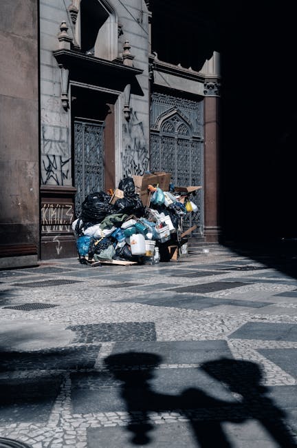 The image depicts an outdoor scene with a large collection of waste materials. Prominently, there are numerous stacked cardboard fruit and vegetable boxes, mainly in red, yellow, and green colors, fea
